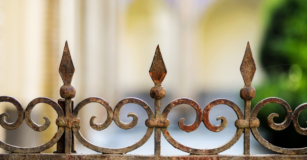 A rusted metal fence with decorative scrollwork and pointed finials stands in front of a blurred house.