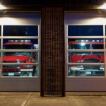 Fire trucks sit inside two illuminated glass garage bays at a fire station while firefighters prepare gear nearby.