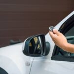 A person presses a garage remote while driving a white truck. A brown garage door is partially open in front of the truck.