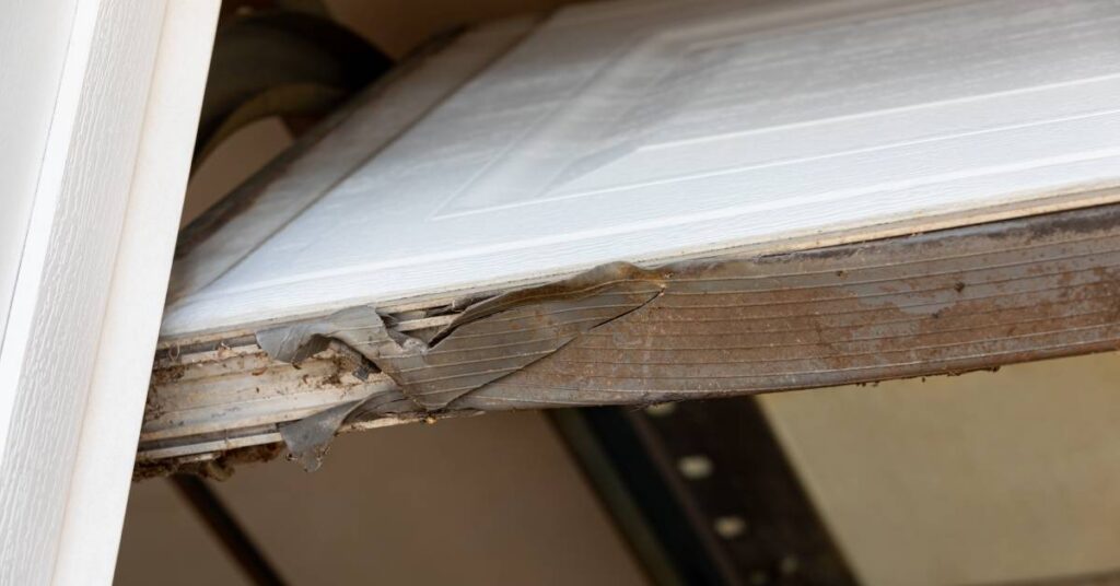 A damaged garage door seal cracks and bends along the bottom edge of a white garage door protecting a residential garage.
