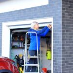 A woman standing on a ladder does maintenance on her garage door with her red car parked in the driveway nearby.