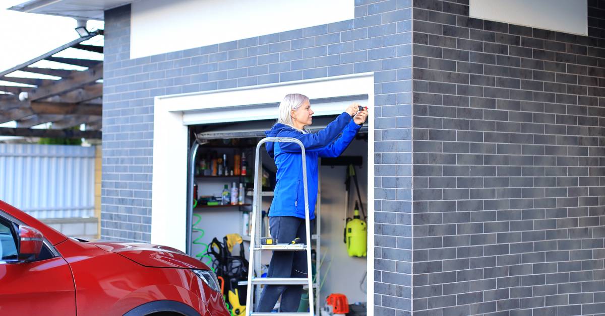 A woman standing on a ladder does maintenance on her garage door with her red car parked in the driveway nearby.