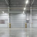 A large rolling steel door is in a bright warehouse interior beneath exposed beams and rows of ceiling lights.