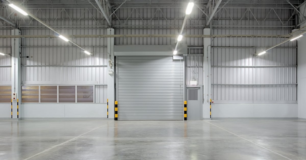 A large rolling steel door is in a bright warehouse interior beneath exposed beams and rows of ceiling lights.