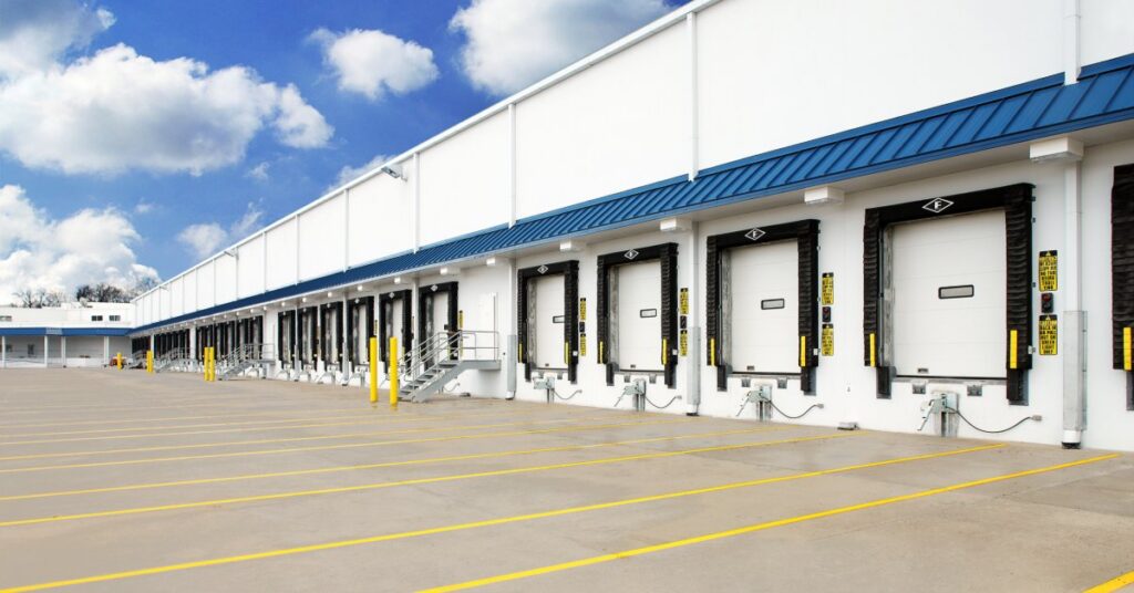 Several loading dock doors line the exterior of a warehouse beneath a blue awning and a bright, cloudy sky.