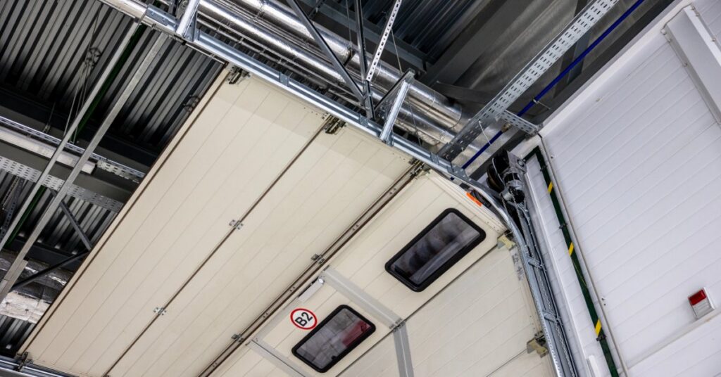 A sectional overhead door curves along ceiling tracks inside an industrial warehouse with exposed ducts and beams.
