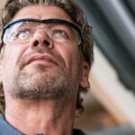 A technician wearing safety glasses inspects a garage door mechanism while looking upward at the overhead hardware.