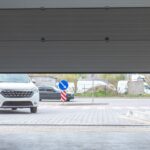 A residential garage door opens as a white car waits in the driveway to pull into the garage and park.