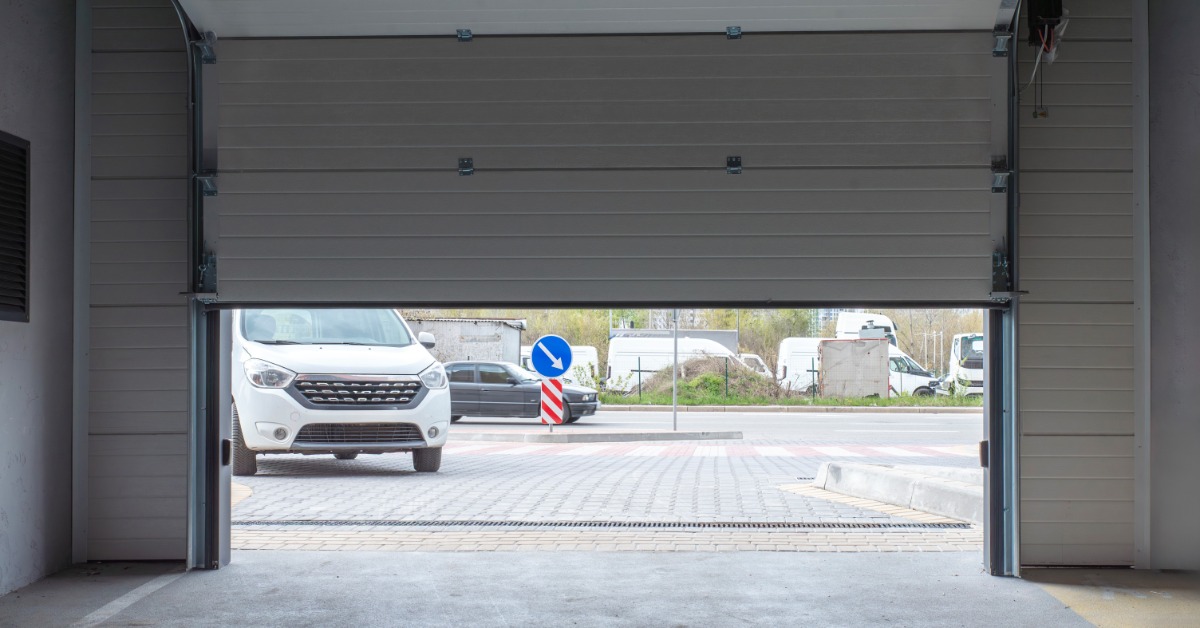 A residential garage door opens as a white car waits in the driveway to pull into the garage and park.