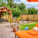 An orange backyard lounge chair and umbrella overlook a fenced backyard patio with a wooden gazebo and green lawn.