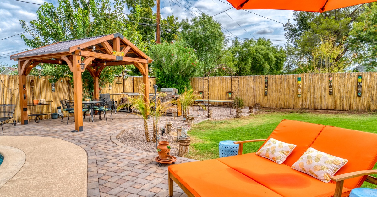 An orange backyard lounge chair and umbrella overlook a fenced backyard patio with a wooden gazebo and green lawn.