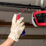 A gloved hand sprays lubricant onto the black chain of a garage door opener hanging from the ceiling.