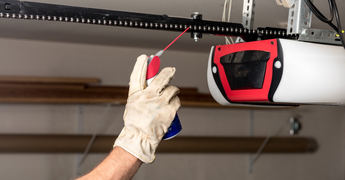 A gloved hand sprays lubricant onto the black chain of a garage door opener hanging from the ceiling.