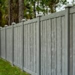 A tall gray privacy fence borders a green lawn as pine trees rise behind the enclosed residential yard.