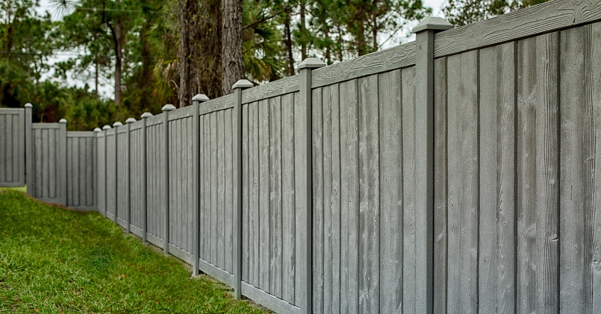 A tall gray privacy fence borders a green lawn as pine trees rise behind the enclosed residential yard.