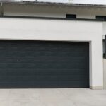 A modern white home frames a wide black garage door beside a clean driveway and a raised garden wall.