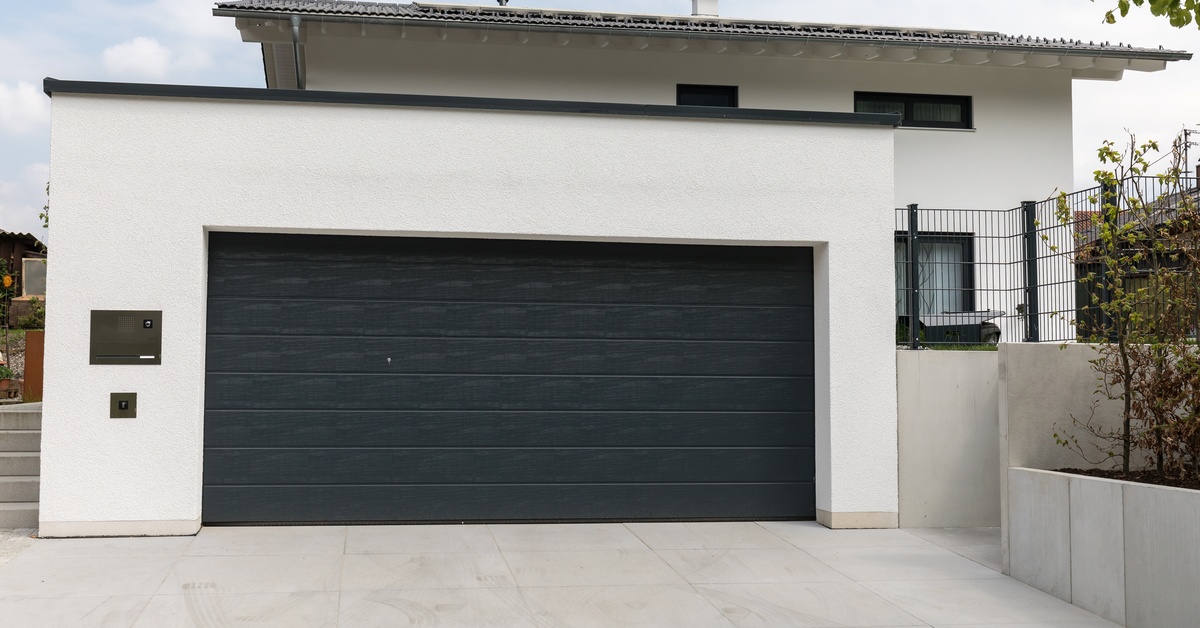 A modern white home frames a wide black garage door beside a clean driveway and a raised garden wall.