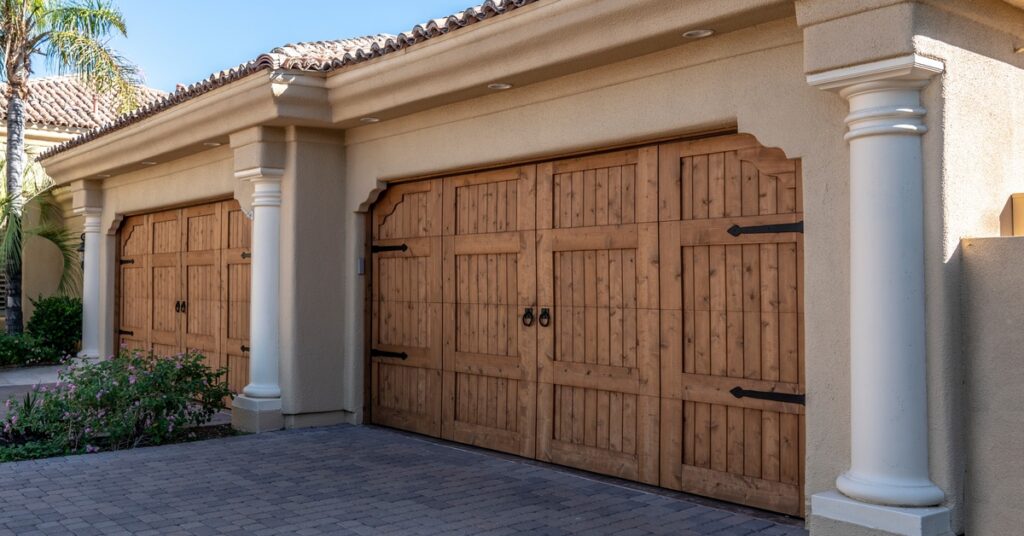 A stucco home features two rustic wooden garage doors beneath a tiled roof and tall decorative columns.
