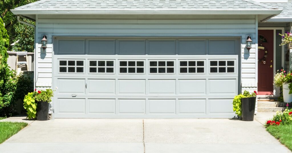 A light blue home features a white paneled garage door with windows, flanked by black lanterns and flower planters.
