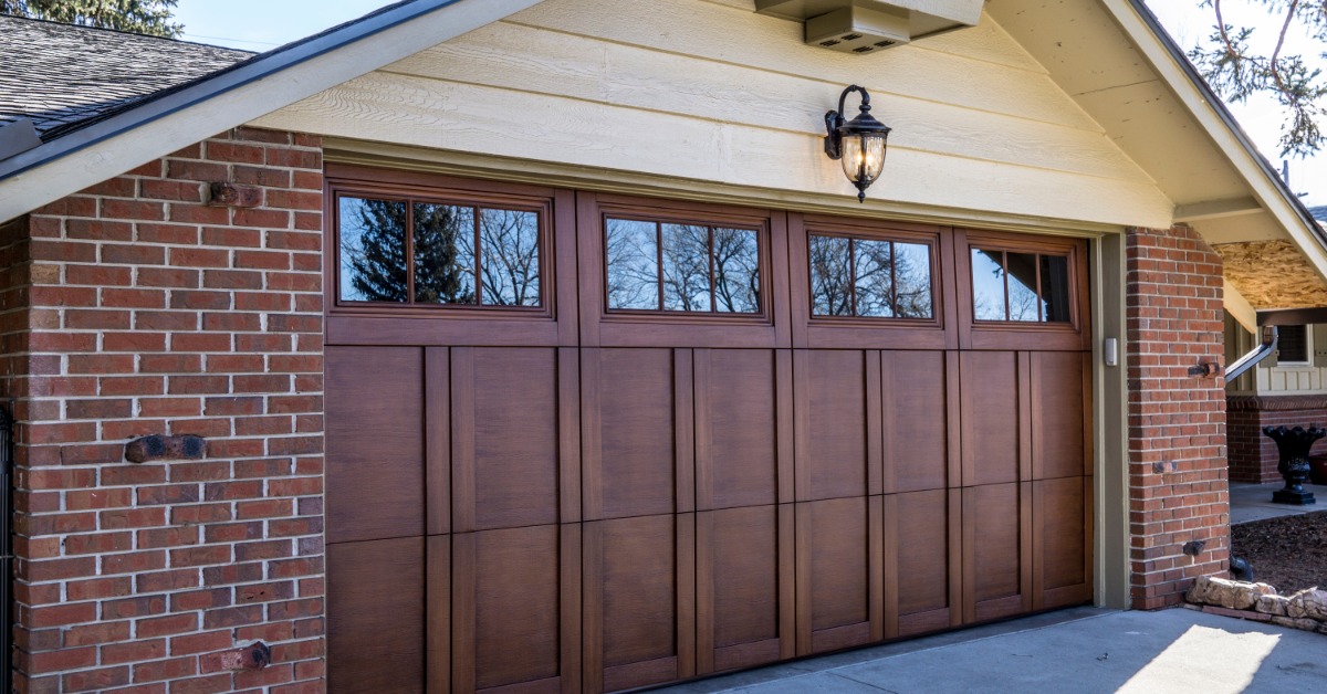 A brick home features a brown wooden garage door with upper windows beneath a beige gable and an exterior lantern.