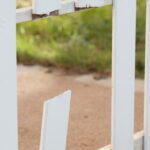A broken white picket fence contains a snapped slat and chipped wood beside a grassy yard and concrete path.