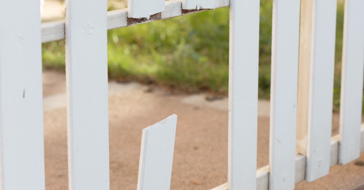 A broken white picket fence contains a snapped slat and chipped wood beside a grassy yard and concrete path.