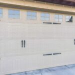 Beige carriage-style garage door with decorative black hinges, handles, and upper windows beneath a wooden pergola.