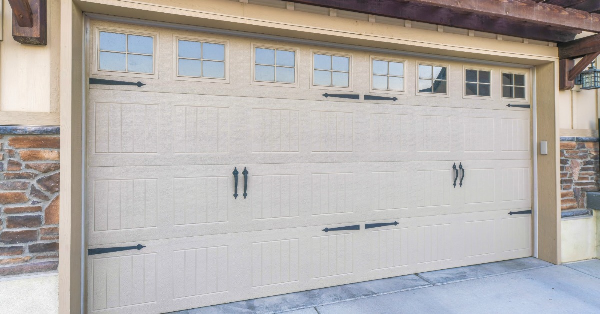Beige carriage-style garage door with decorative black hinges, handles, and upper windows beneath a wooden pergola.