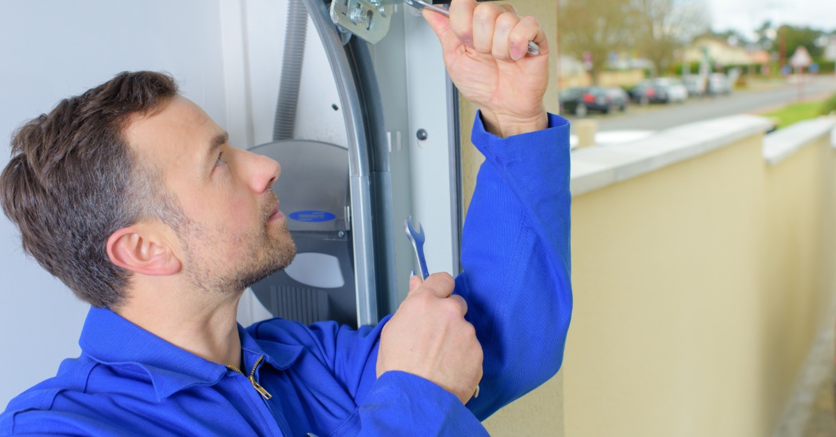 Garage door technician in blue workwear using a wrench to adjust the metal hardware of an overhead garage door.